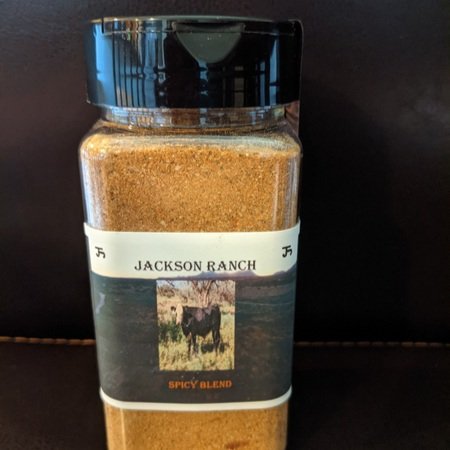 Container of Jackson Ranch Spicy Blend seasoning, featuring a clear jar with a black lid and a label depicting a cow and scenic ranch background. Ideal for enhancing the flavor of various dishes.