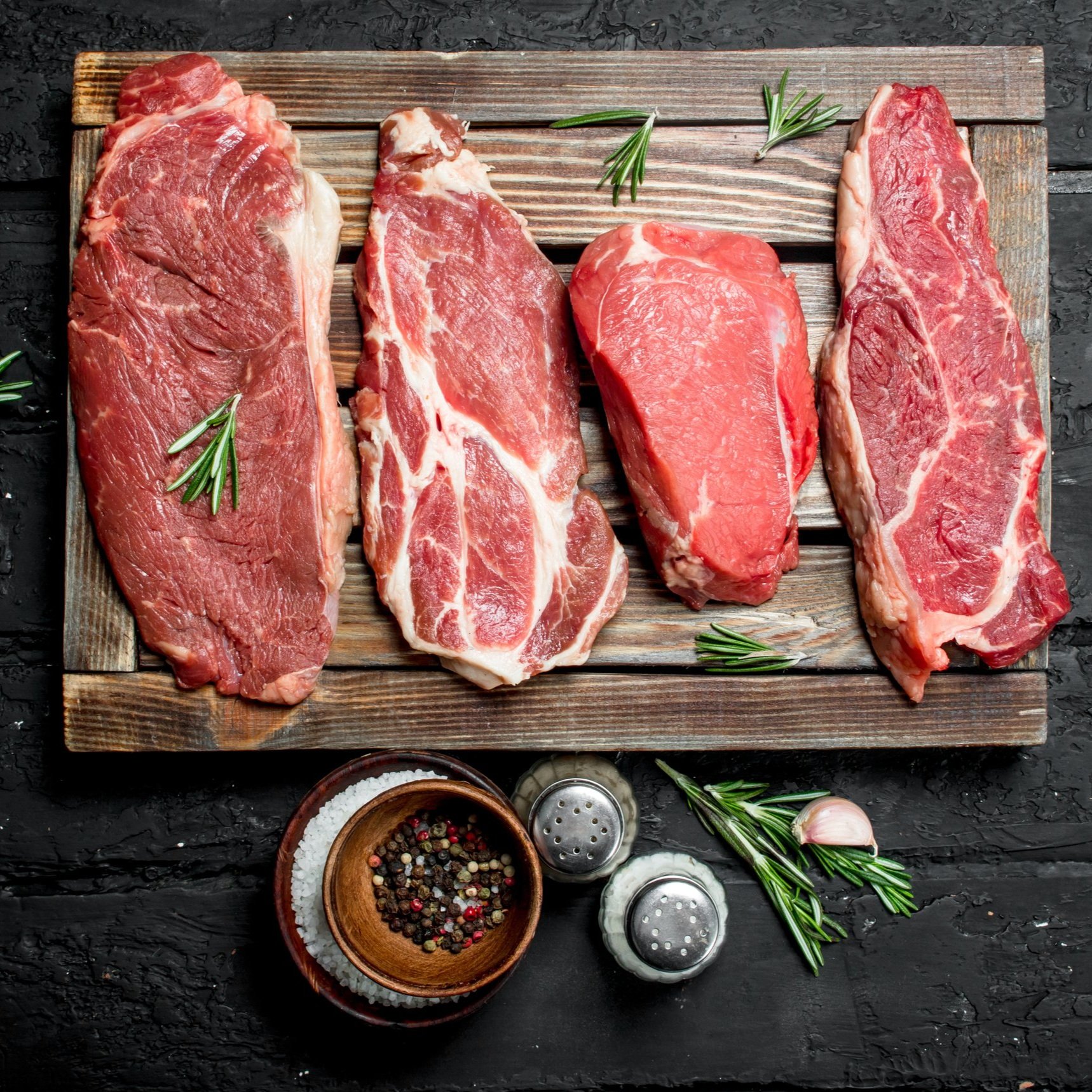 Assorted cuts of raw beef steaks displayed on a wooden cutting board, garnished with fresh rosemary, alongside a bowl of mixed peppercorns and salt shakers, set against a dark textured background.