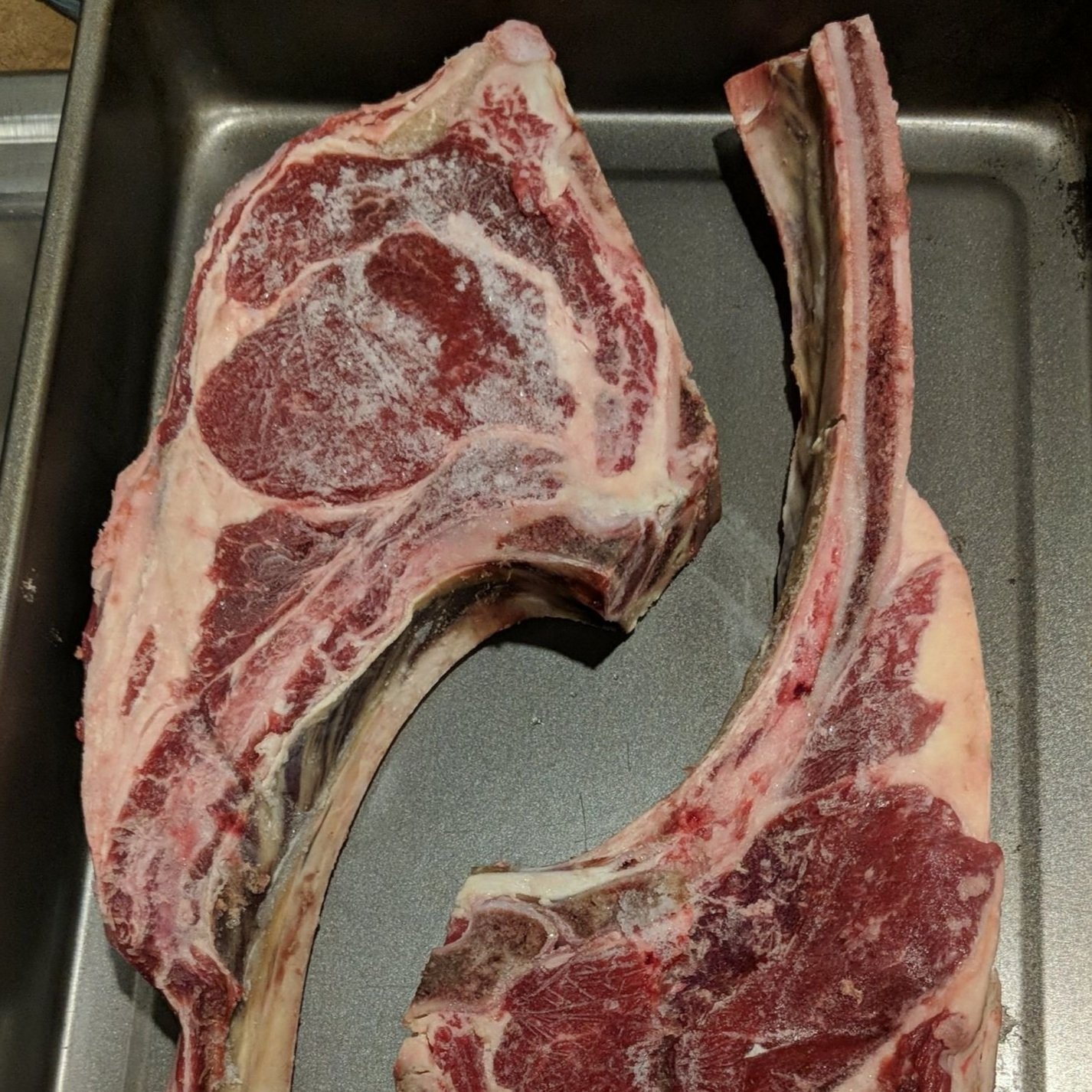 Two large, frozen tomahawk steaks displayed on a gray baking tray, showcasing their marbled red meat and white fat.