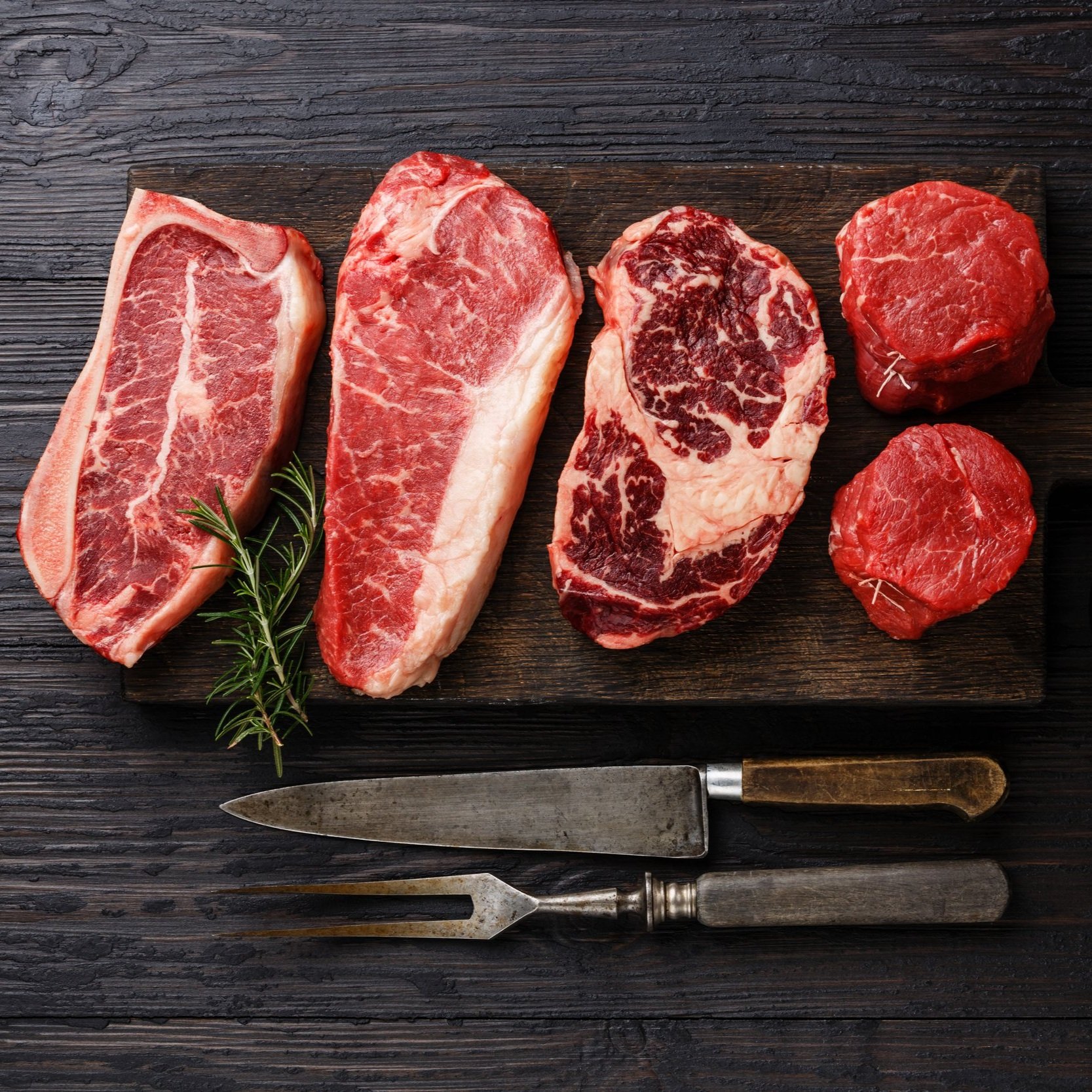 Assorted cuts of raw beef, including ribeye and tenderloin, displayed on a wooden cutting board with a sprig of rosemary and stainless steel knives. Ideal for culinary presentations and meat preparation ideas.